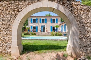 A stone archway framing a building with a pool and a well-kept lawn. - Beautiful stone property with view and heated pool, 6 km from Gordes (Murs)