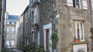A narrow cobblestone street lined with stone buildings, some with potted plants on the ground floor.