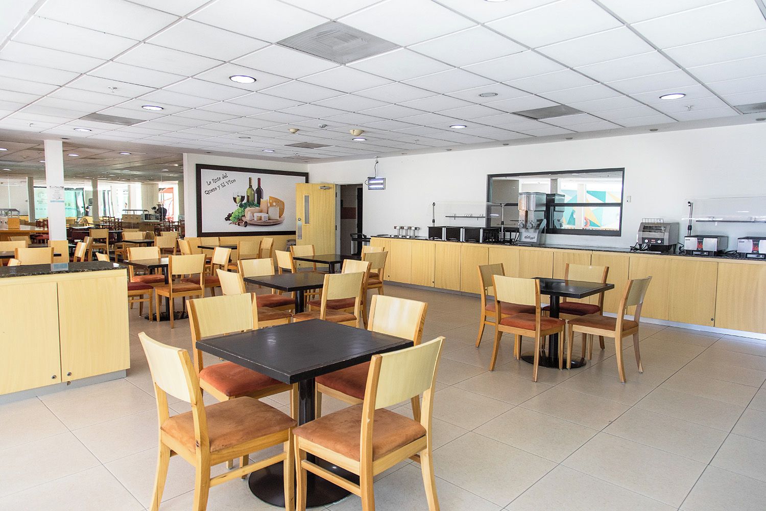 A spacious cafeteria with multiple tables and chairs, a kitchen area with appliances, and a large poster on the wall.
