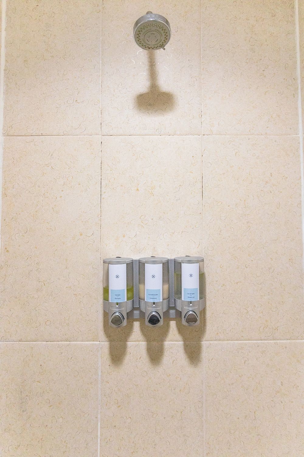 A bathroom with a showerhead and three wall-mounted dispensers for shampoo, conditioner, and body wash.
