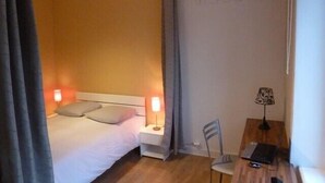 Une chambre d’hôtel avec un lit, deux lampes de chevet, un bureau avec une chaise et un mur sur lequel on peut apercevoir partiellement un nom.