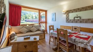 A cozy room with a wooden sofa, dining table, and chairs. A window with red curtains offers a view of trees and a building. - Apartment 4/5 PERS ARCS 1800 - WIFI (Arc 1800)