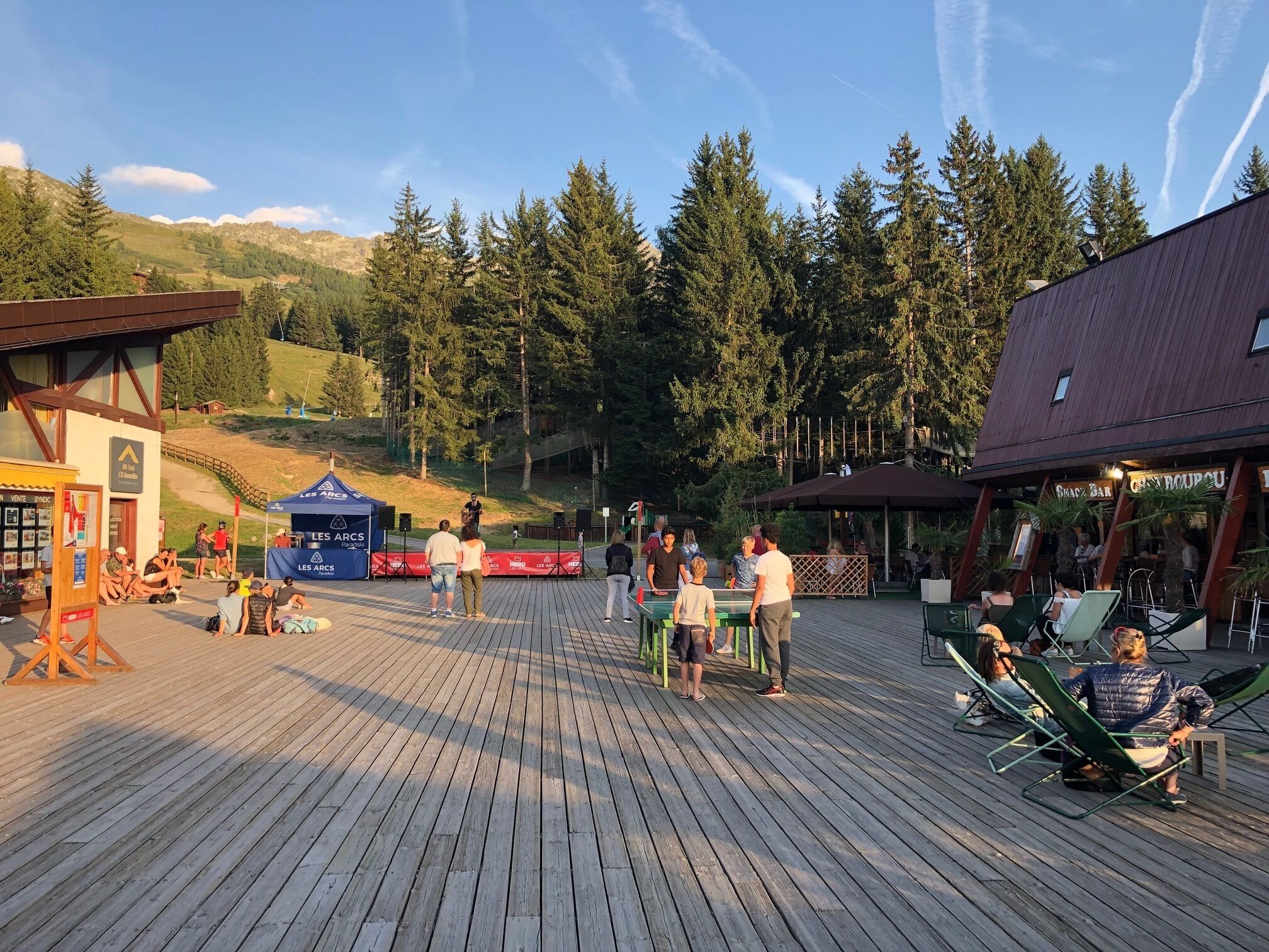 Una terraza de madera con gente sentada y de pie, un escenario con una carpa azul y un edificio con un letrero que dice "LES ARCS".