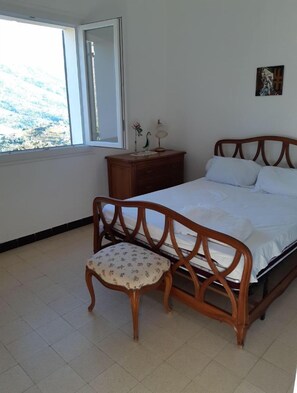 Ein Schlafzimmer mit einem hölzernen Bett, einem Nachttisch, einem Stuhl und einem Fenster mit Blick auf eine gebirgige Landschaft.