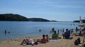 A beach with people sunbathing and swimming, a sailboat, and a forested hill in the background. - Country house 4pers (Montboudif)