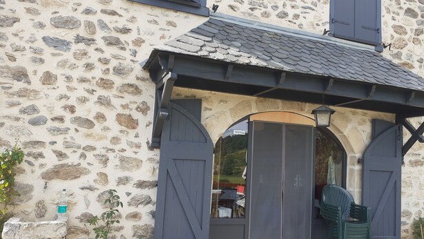 A stone building with a tiled roof, a wooden door, and a small outdoor seating area with a chair and a table. - Country house 4pers (Montboudif)