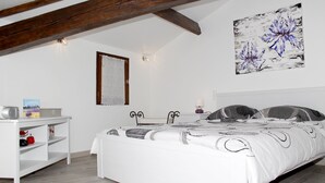 Un dormitori amb un llit blanc, una tauleta de nit blanca, una estant amb objectes decoratius, un quadre a la paret i bigues de fusta al descobert.
