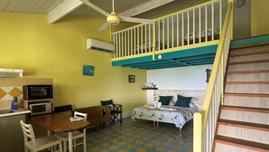 A room with a checkered floor, a dining table with chairs, a bed, a staircase, and a ceiling fan.