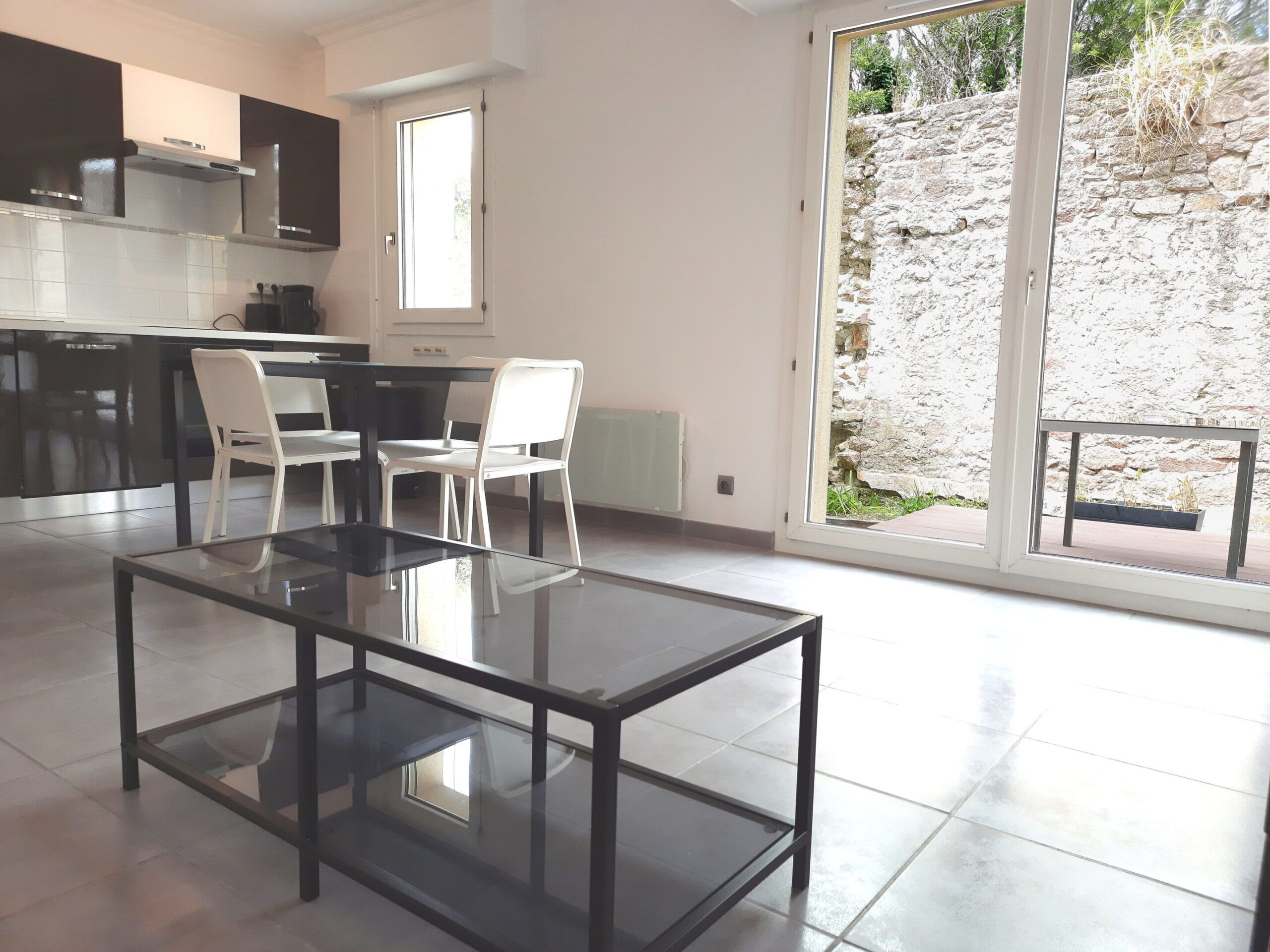 A modern kitchen with a dining area, a glass table, and a view of a stone wall outside.