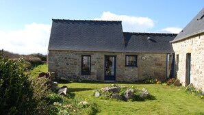 Une maison en pierre avec un toit sombre, une porte d’entrée et un petit jardin agrémenté de rochers et de fleurs.