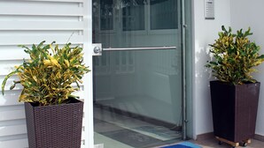 L’entrée d’un hôtel, avec une porte en verre, un paillasson bleu et deux plantes en pot.