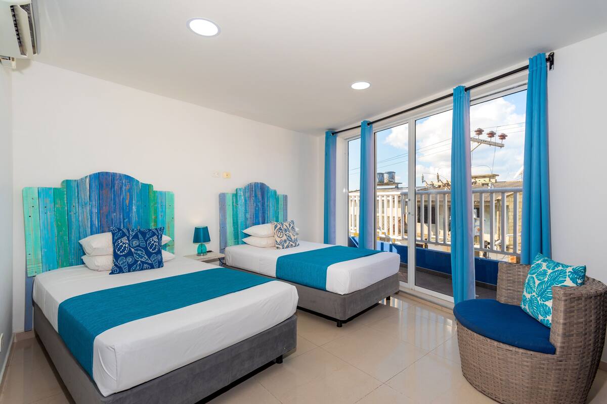 Una habitación de hotel con dos camas, un balcón con cortinas azules y una silla de mimbre.