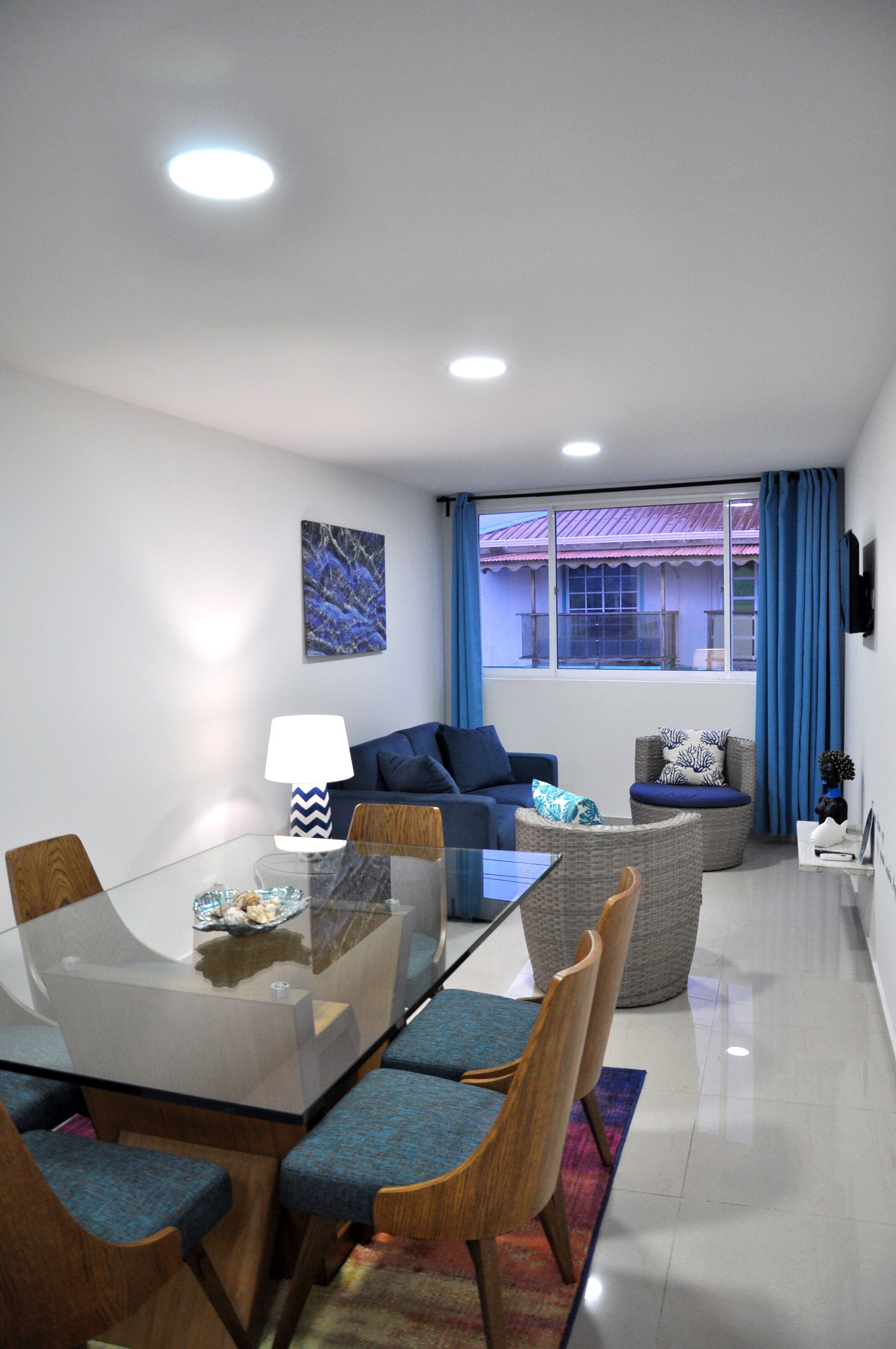 A modern living room with a glass dining table, blue upholstered chairs, a sofa, and a wicker armchair.
