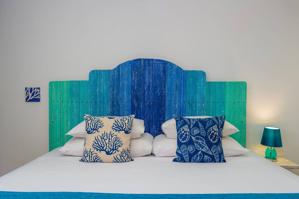Una cama con cabecera azul, dos almohadas decorativas y una lámpara turquesa sobre una mesita de noche.
