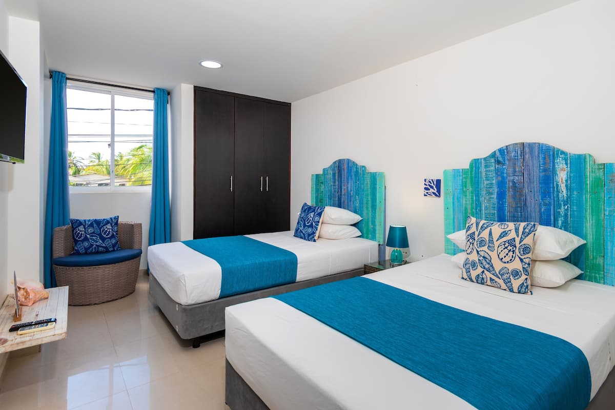 Habitación de hotel con dos camas, televisión, ventana con cortinas y vista a palmeras.