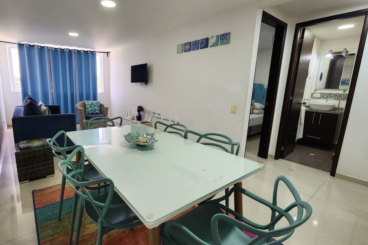 Un comedor con mesa y sillas, un sofá, un televisor y una cocina visible a través de una puerta abierta.