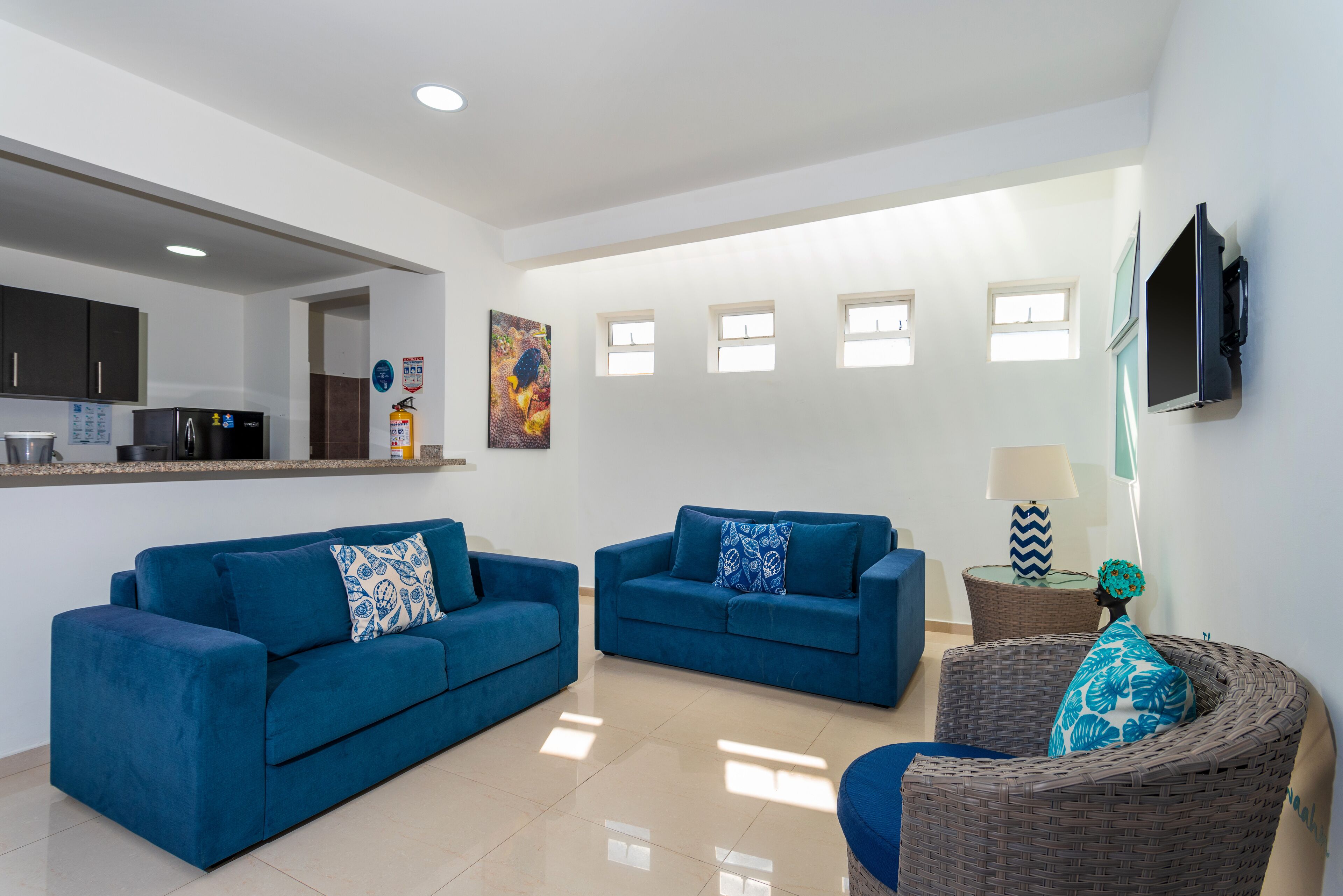 A modern living room with two blue sofas, a wicker chair, a flat-screen TV mounted on the wall, and a kitchen area visible through an open doorway.