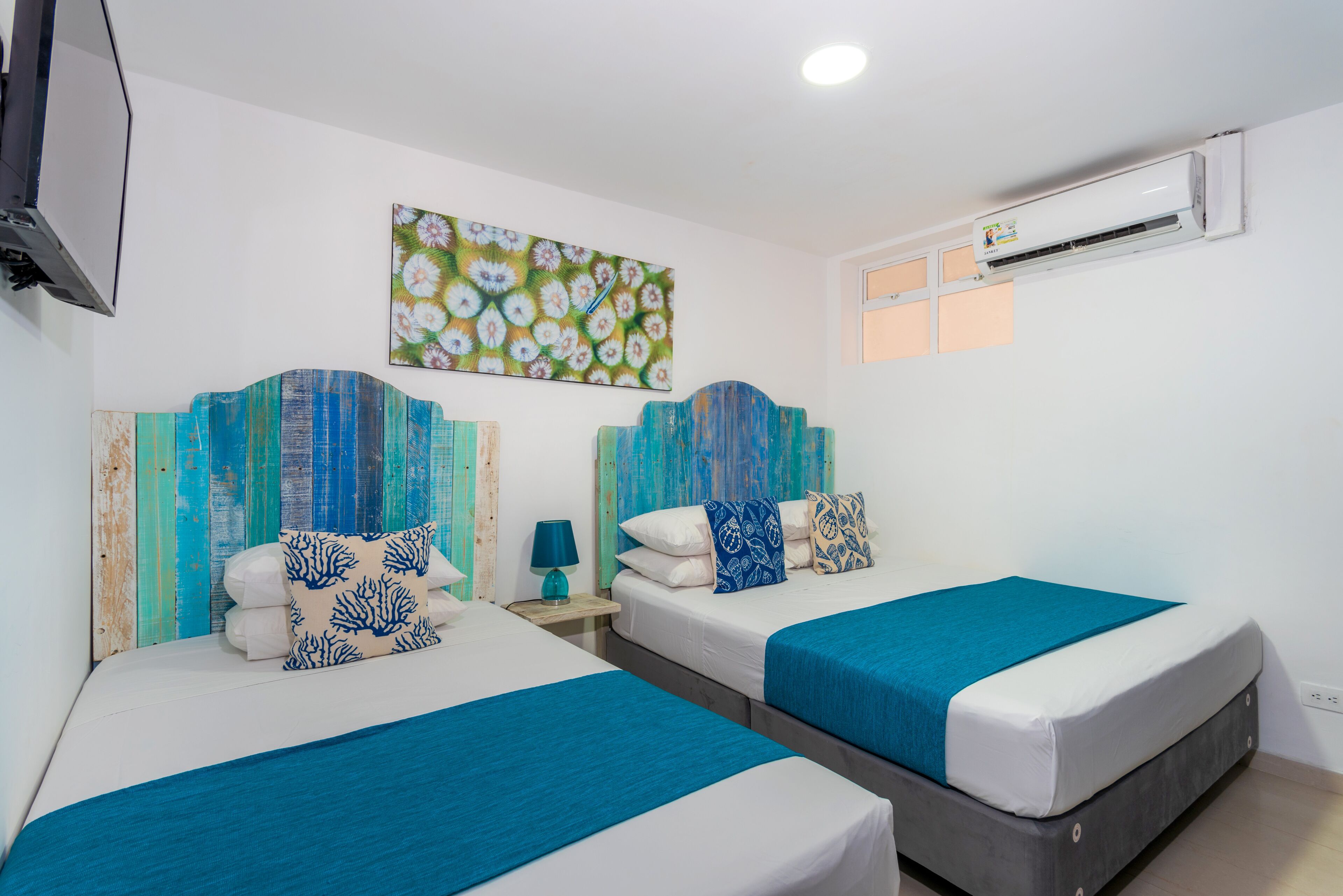 Habitació d'hotel amb dos llits, cadascun amb una cobra blava i almoinades decoratives. Hi ha un quadre a la paret, una taula petita amb una làmpada i una unitat de climatització penjada a la paret.