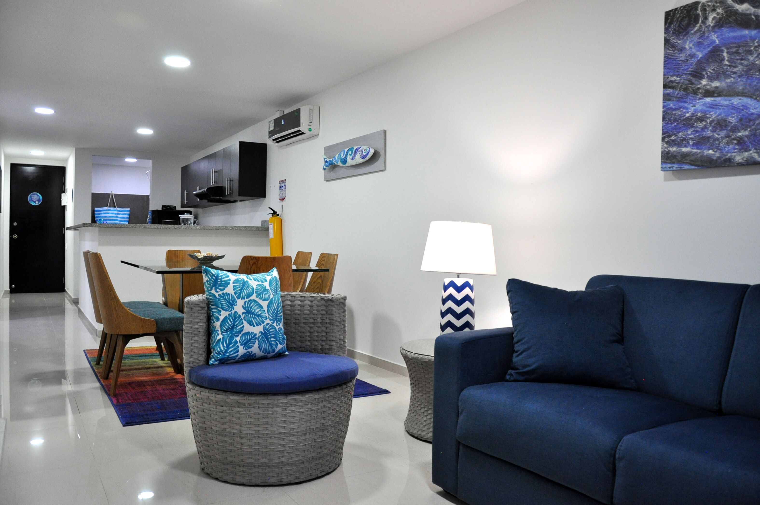 A modern living room with a blue sofa, wicker chairs, a dining area with a table and chairs, and a kitchen in the background.