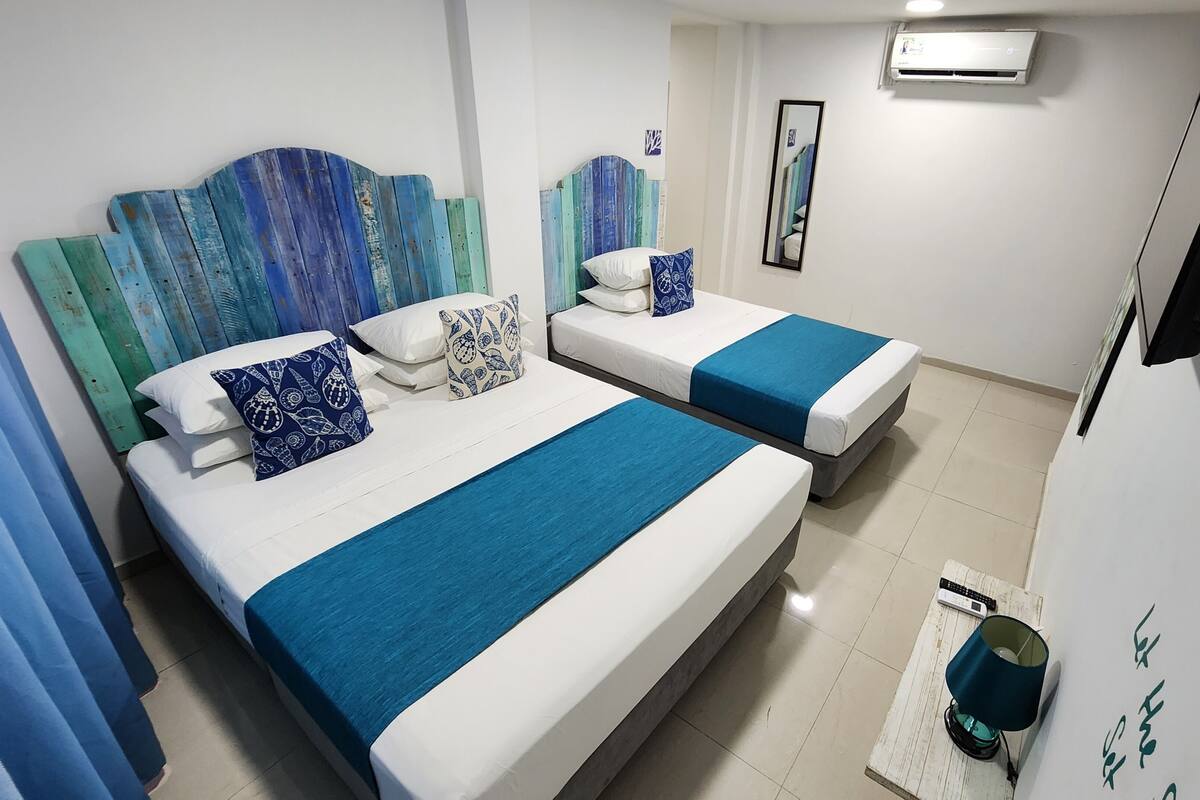 Una habitación de hotel con dos camas, cada una con ropa de cama azul y blanca, un televisor, aire acondicionado y una mesita de noche con una lámpara.