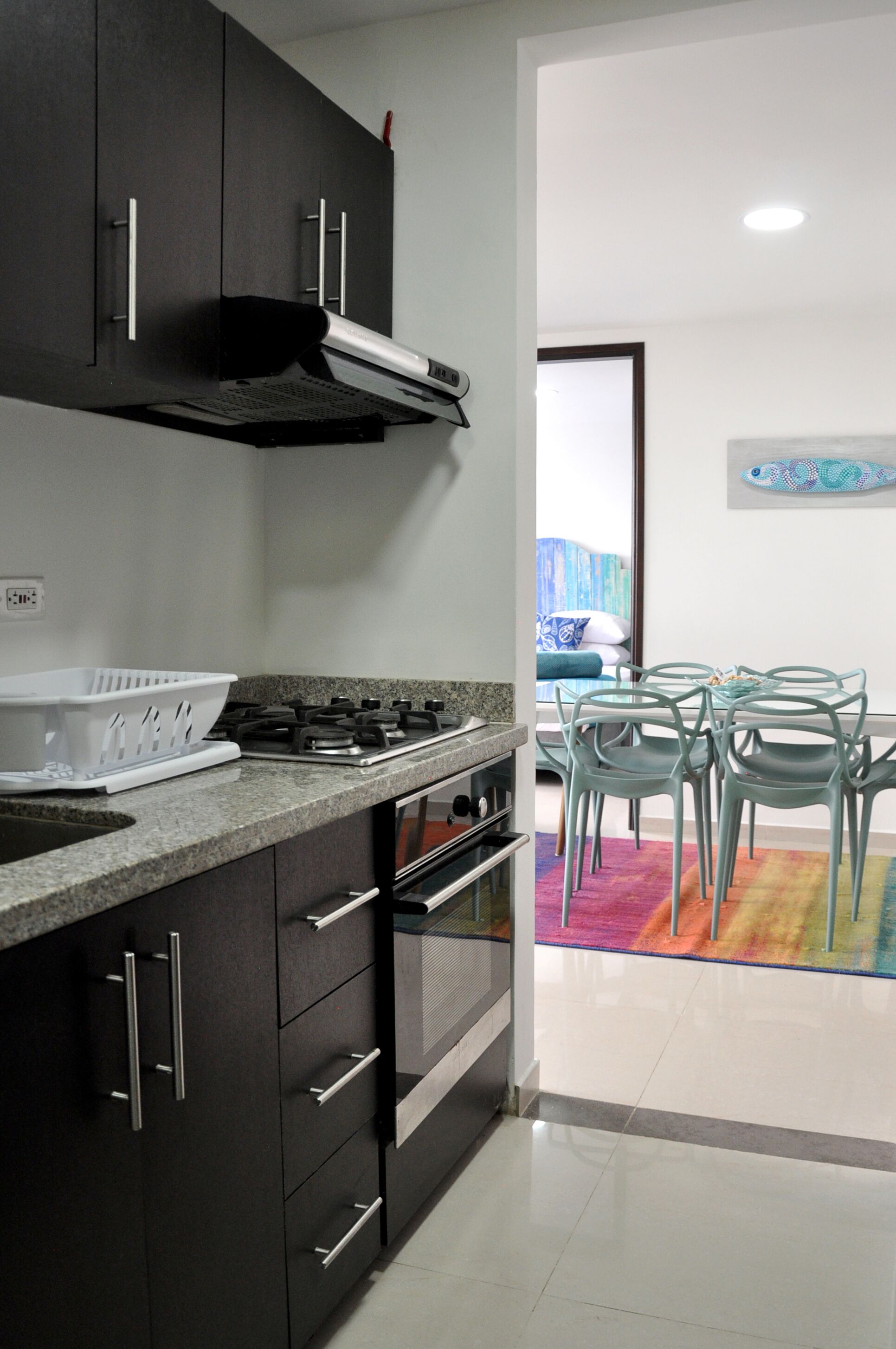 A modern kitchen with dark cabinets, a gas stove, and a dining area with a colorful rug and glass chairs.
