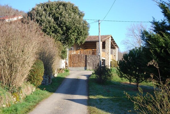 A rural road leading to a stone house with a wooden gate, surrounded by trees and shrubs.