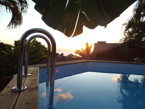 Piscina com escada de metal, rodeada por palmeiras e um edifício ao fundo.