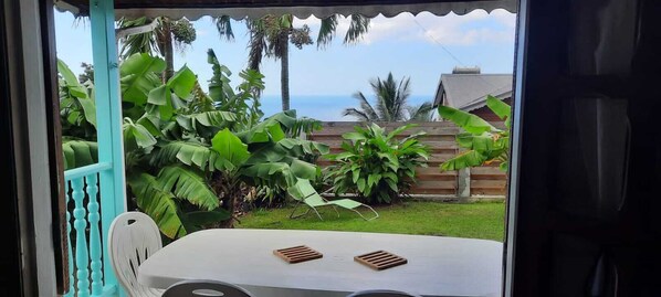 Vista da janela com uma mesa e cadeiras, cerca de madeira e plantas verdes exuberantes.