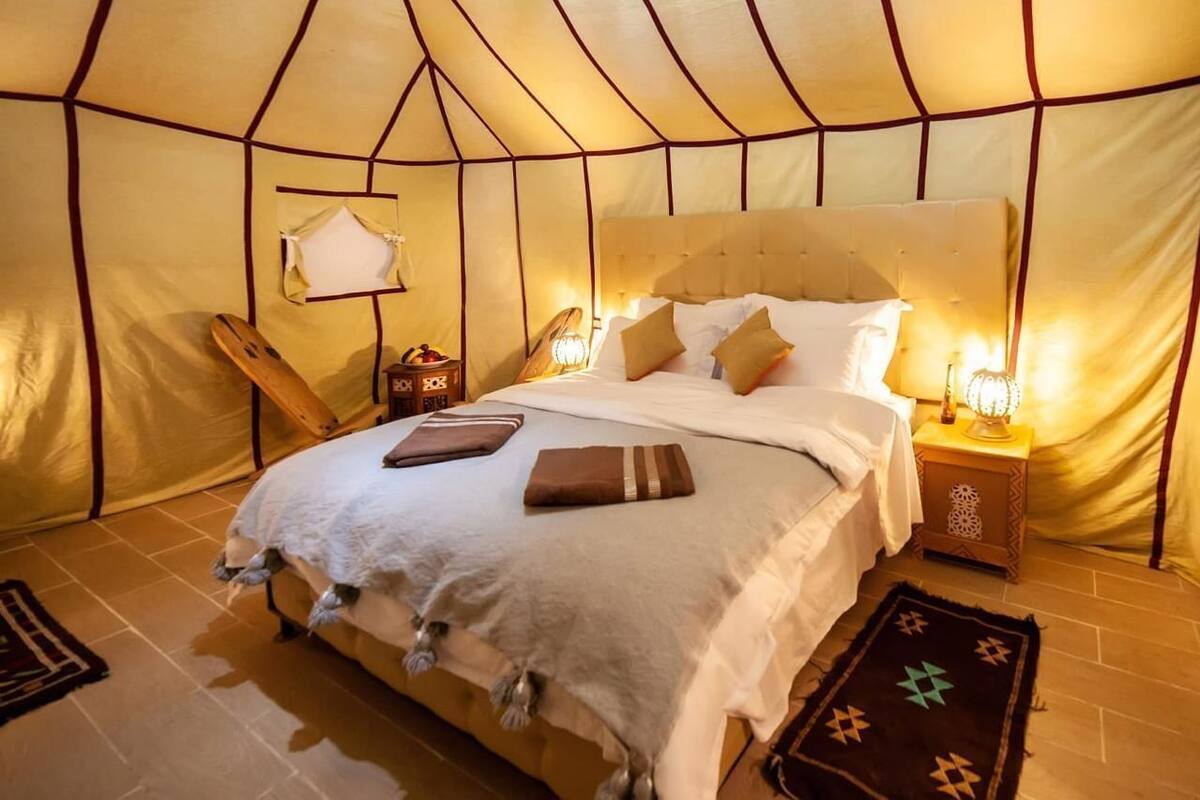 A tent-style room with a bed, pillows, a nightstand with a lamp, and a small rug.