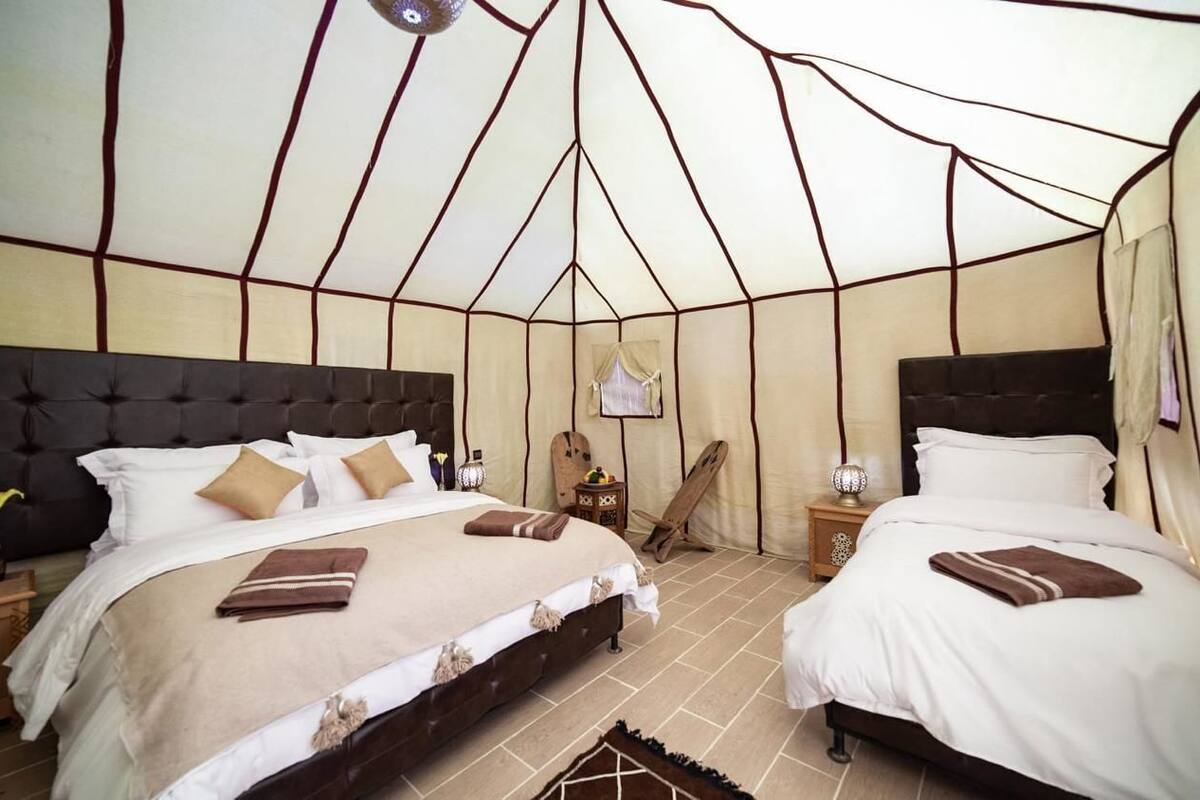 A tent-style room with two beds, a wooden chair, and a small table with a lamp.