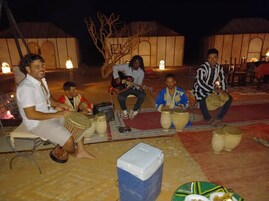 Un grupo de personas sentadas en el suelo tocando tambores tradicionales en un espacio al aire libre, con tiendas de campaña al fondo.