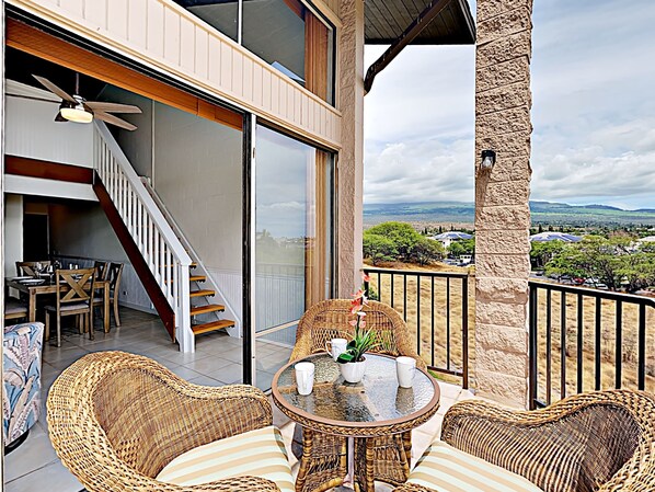 A balcony with wicker furniture, a dining area with a table and chairs, and a view of a landscape with trees and buildings.
