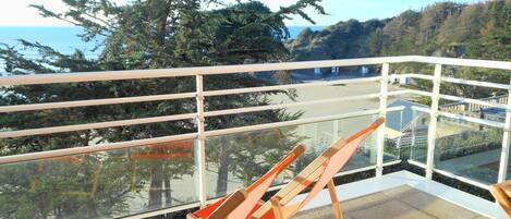 Ein Balkon mit Glasgeländer, zwei orangefarbene Stühle und Blick auf das Meer und die Hügel.