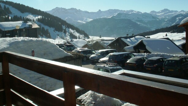 Un pueblo montañoso nevado con una valla de madera en primer plano, un estacionamiento con coches y edificios cubiertos de nieve.