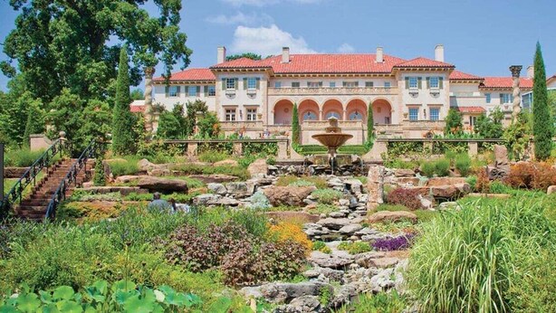 Un grand bâtiment à plusieurs étages, doté d’un toit recouvert de tuiles rouges, entouré de jardins luxuriants et agrémenté d’une fontaine centrale.