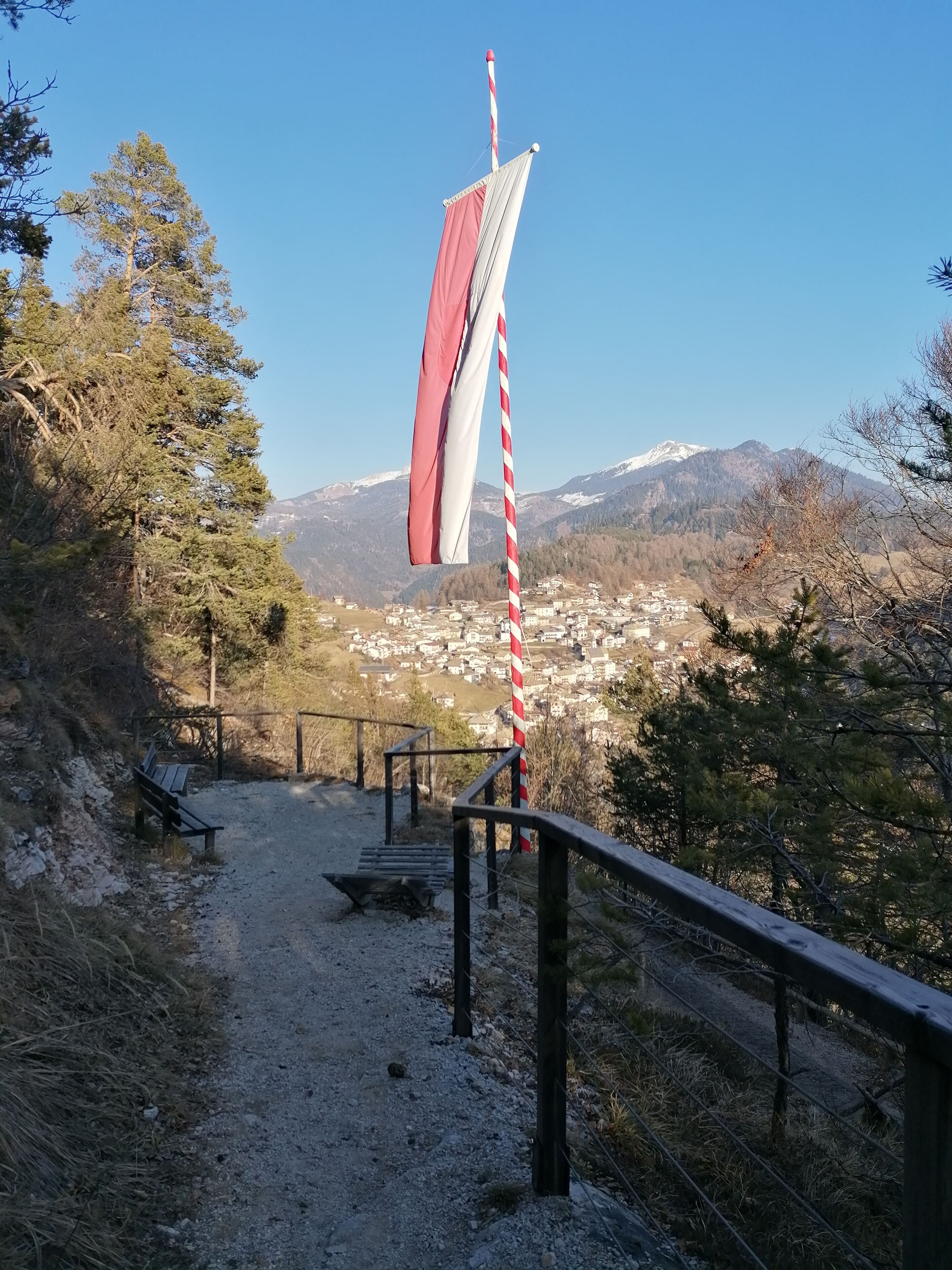 A flagpole with a flag, a wooden bench, a dirt path, and a view of a town and mountains in the background.