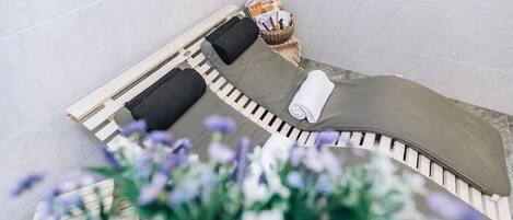 A spa room with a curved bench, two black cushions, a white towel, and a basket of magazines.