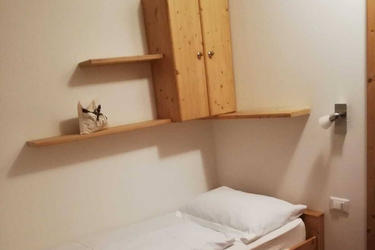 A small, single-bed room with a wooden bed frame, a wooden wardrobe, and a small shelf above the bed.