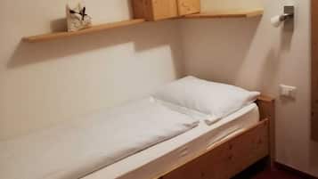A small, single-bed room with a wooden bed frame, a wooden wardrobe, and a small shelf above the bed.