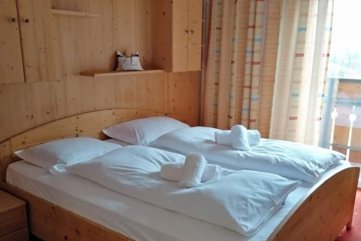 A wooden bed with white linens and towels, a wooden nightstand, a shelf with a bag, and a window with curtains.