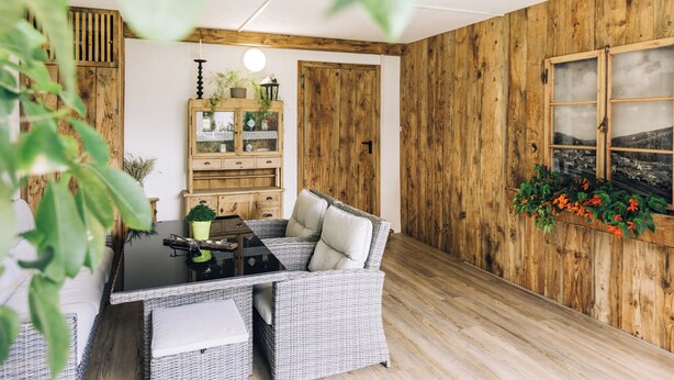 Ein Zimmer mit einer Holzwand, einem Korbsessel, einer Holzkommode mit Pflanzen und einem Fenster mit Blick.