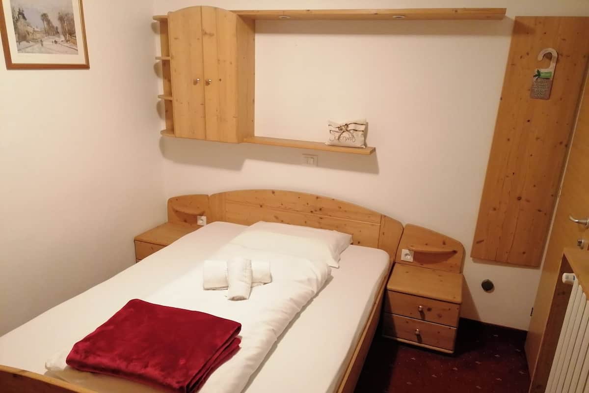 A single bed with a red blanket, wooden headboard, bedside tables, a wooden wardrobe, and a small shelf with a decorative item.