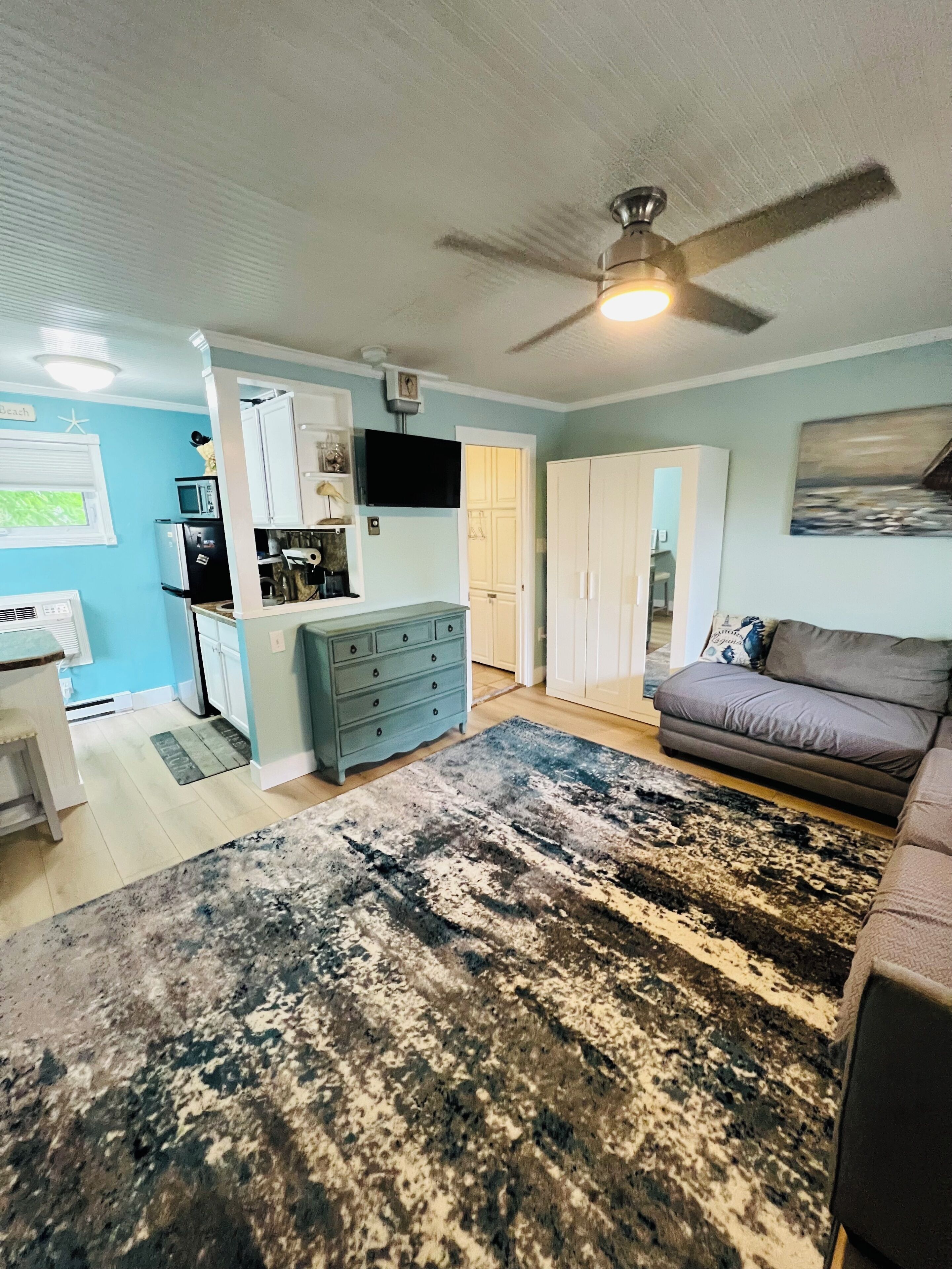 A living room with a ceiling fan, a grey sofa, a large rug, and a kitchen area with cabinets and appliances.