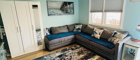 A room with a ceiling fan, a large sectional sofa, a painting of a pier, a window with blinds, and a small table.