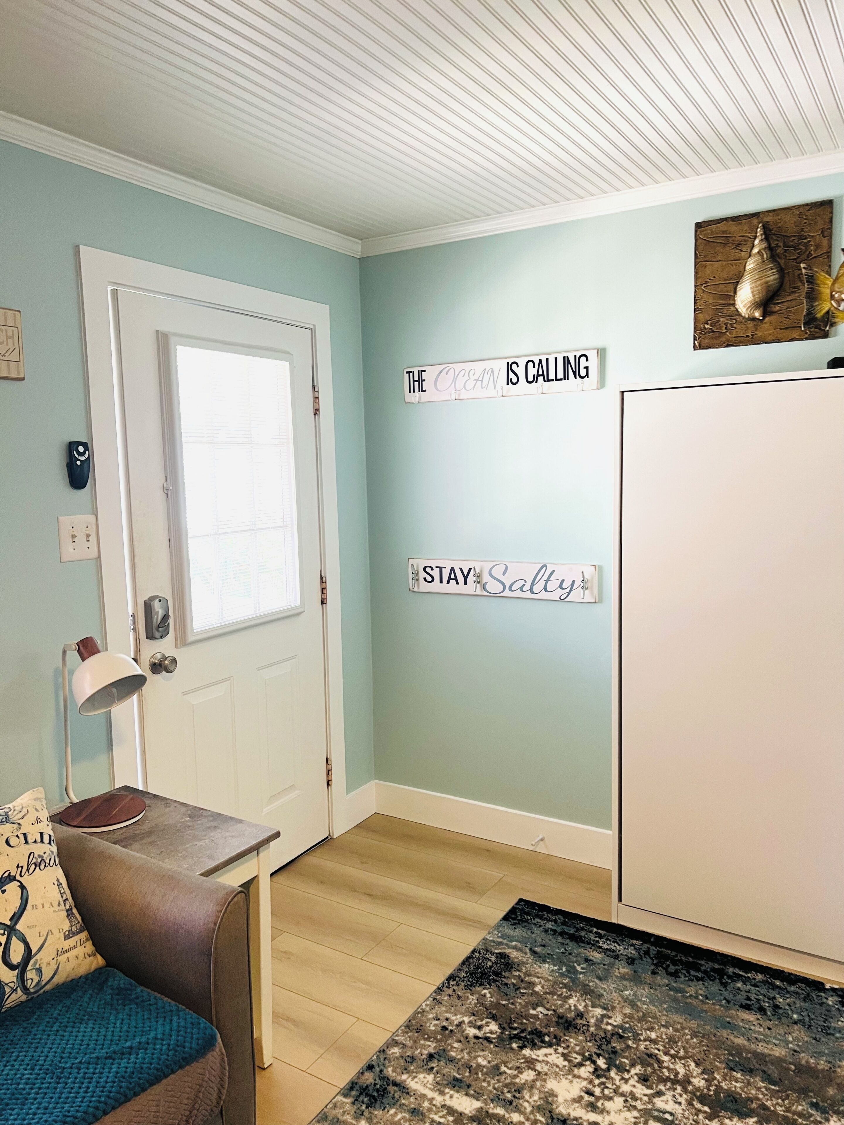 A room with a door, a wall-mounted sign reading 'THE OCEAN IS CALLING,' a small table with a lamp, and a rug.