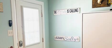 A room with a door, a wall-mounted sign reading 'THE OCEAN IS CALLING,' a small table with a lamp, and a rug.