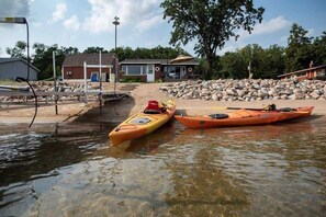 Kayaks amarrados cerca de un edificio con un muelle, un cobertizo pequeño y un montón de rocas al fondo.