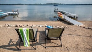 A lakeside view with a dock extending into calm water, two chairs with towels, and a clear sky.