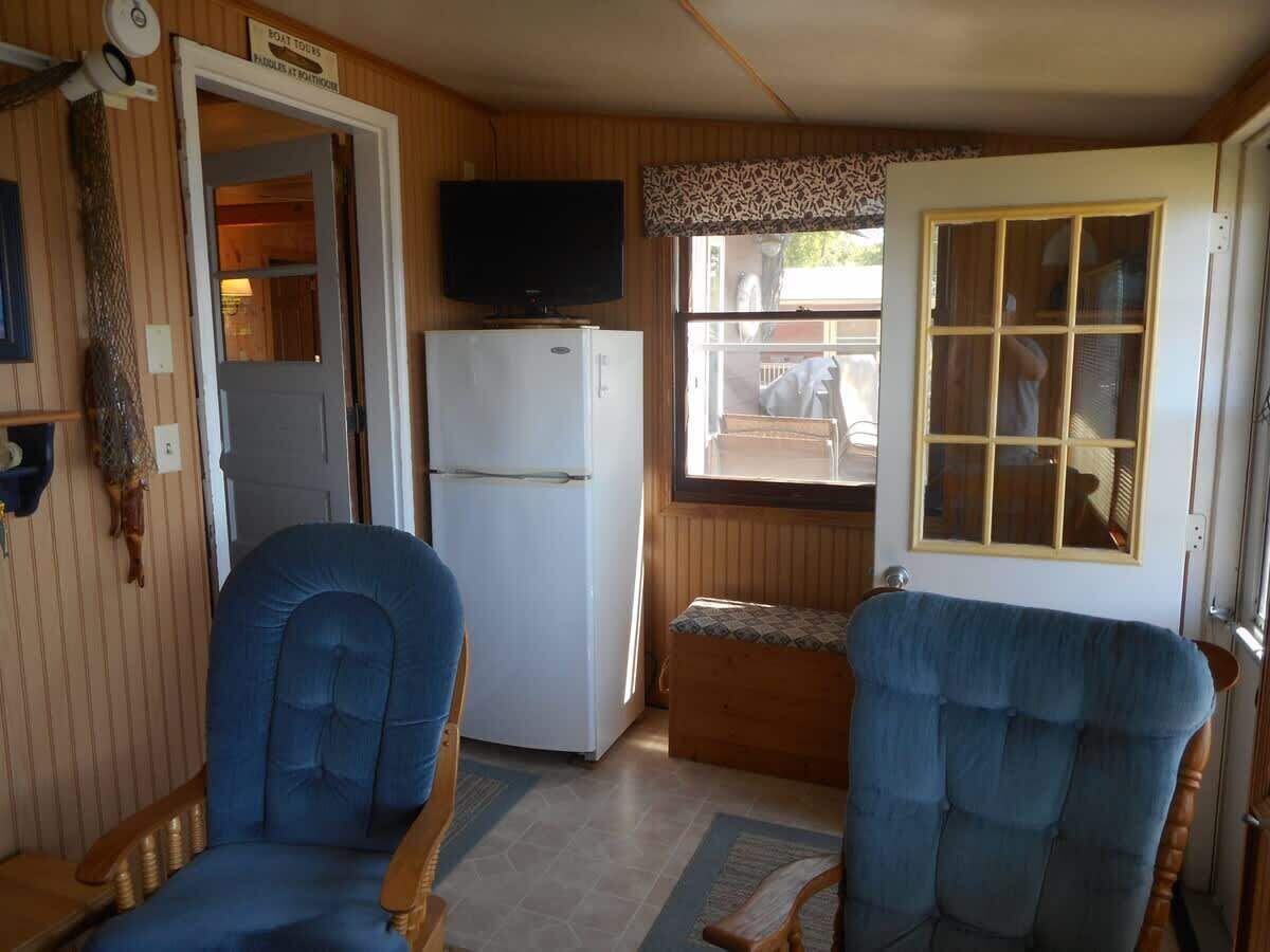 Ein gemütlicher Raum mit Fernseher, Kühlschrank, zwei blauen Stühlen und einem Fenster mit Ausblick nach draußen.