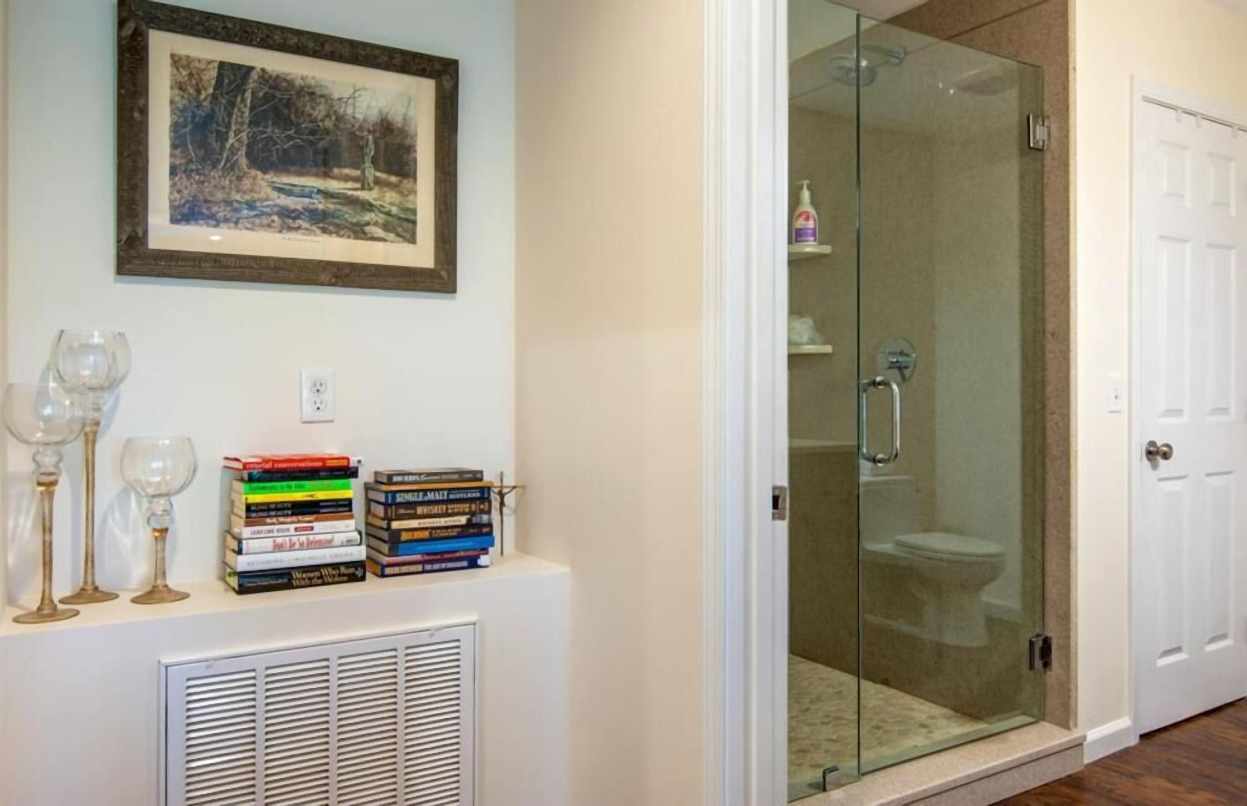 Un baño con cabina de ducha de vidrio, una puerta blanca y una repisa con libros y cristalería.